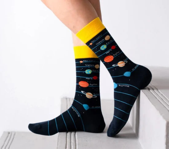 A pair of crew socks with a galaxy and star pattern, featuring planets and stars on a black background. The socks are positioned on a set of stairs with one foot raised, displaying the design and fit.
