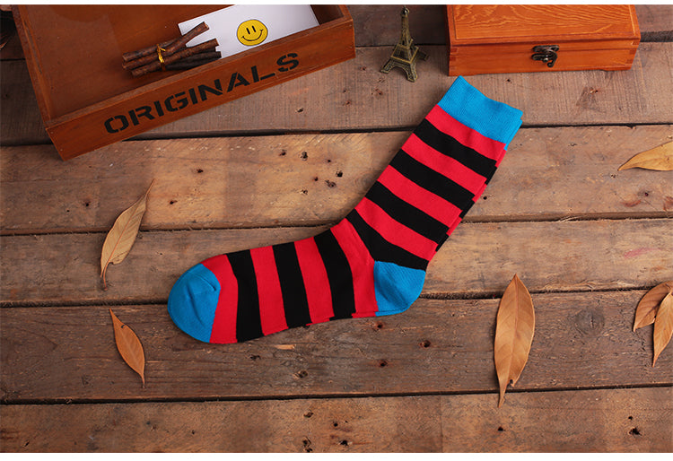 A pair of striped crew socks in red, blue, and black, displayed on a wooden surface with a box of cigars and leaves scattered around.